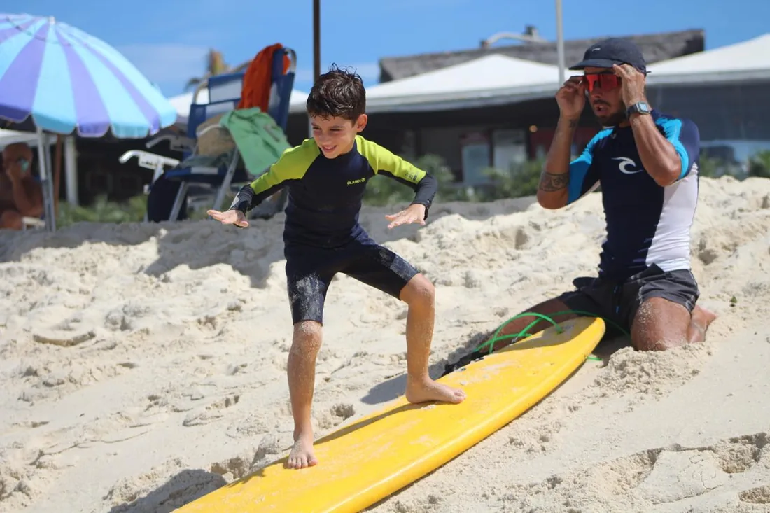 Aluno treinando a base do surf na areia com instrutor ao lado